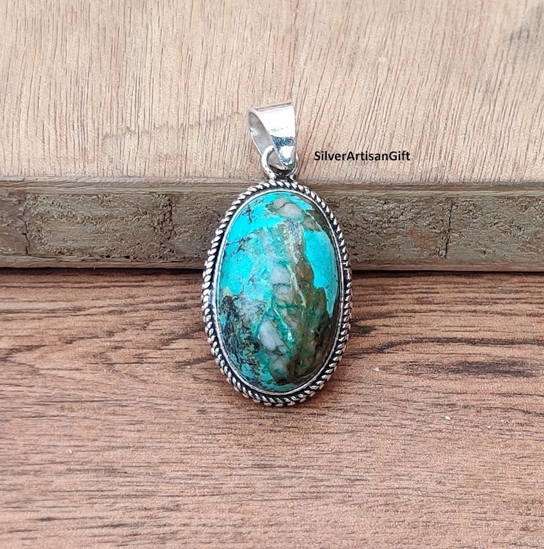 May include: Oval turquoise pendant with a silver setting. The turquoise is a vibrant blue-green color with white and brown marbling. The pendant is on a wooden surface.