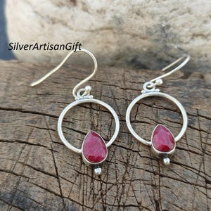 May include: A pair of silver earrings with a red gemstone drop hanging from a circle. The earrings have a hook fastening.