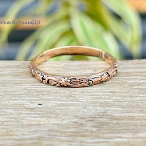 May include: A rose gold ring with a floral pattern. The ring has intricate carvings of flowers and leaves around the band. The ring is displayed on a wooden surface with a blurred green background.