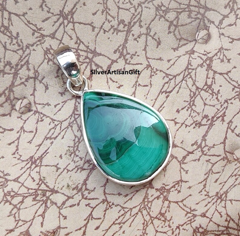 May include: A teardrop-shaped pendant with a polished, vibrant green malachite stone set in a silver bezel. The stone displays natural swirling patterns. The pendant has a silver bail for attaching to a chain. The text "SilverArtisanGift" is visible.