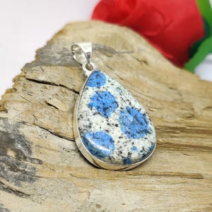 Peut inclure: Un pendentif en argent avec une pierre bleu et blanc en forme de larme avec un motif moucheté. Le pendentif est sur une surface en bois.