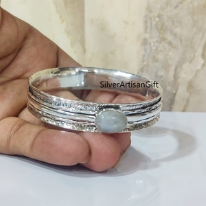 May include: Silver bangle bracelet featuring a round, light grey gemstone. The bracelet has a hammered texture and is made of three bands.