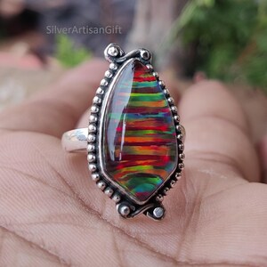 Puede incluir: Un anillo de plata con una piedra preciosa grande en forma de l&aacute;grima con bandas vibrantes de rojo, naranja, amarillo y verde. La piedra preciosa est&aacute; engastada en un bisel de plata detallado con un borde de cuentas. El texto "SilverArtisanGift" es visible.