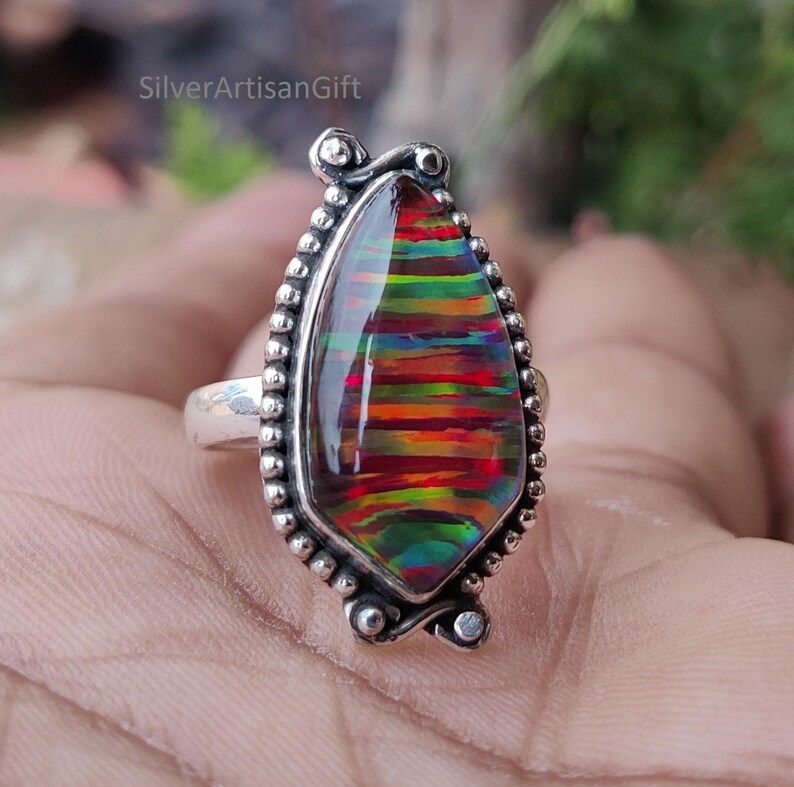 Puede incluir: Un anillo de plata con una piedra preciosa grande en forma de l&aacute;grima con bandas vibrantes de rojo, naranja, amarillo y verde. La piedra preciosa est&aacute; engastada en un bisel de plata detallado con un borde de cuentas. El texto "SilverArtisanGift" es visible.
