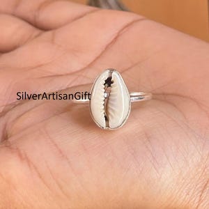 May include: A silver ring featuring a white and tan cowrie shell. The shell is oval-shaped and set in a simple silver band. The text "SilverArtisanGift" is visible on the image.