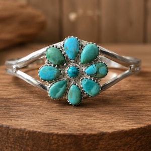 May include: A silver cuff bracelet featuring a central flower-shaped cluster of eight pear-shaped turquoise stones surrounding a single round turquoise stone. The metal band splits into two arms on each side of the floral centerpiece.