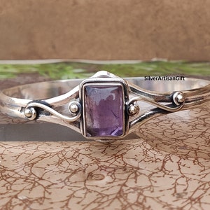 May include: Silver bangle bracelet featuring a purple amethyst gemstone set in a rectangular frame. The bracelet has a delicate, curved design with small silver beads.