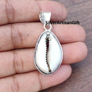 Puede incluir: Un colgante de plata con una concha de cauri blanca. La concha está engastada en un marco de plata y tiene un acabado liso y pulido. El colgante es perfecto para añadir un toque de belleza natural a cualquier atuendo.