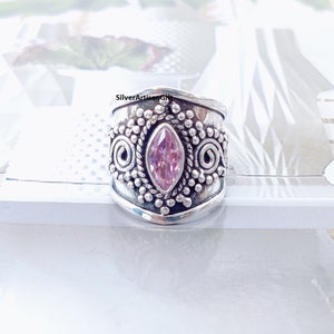 May include: A silver ring with a pink gemstone set in a decorative design. The ring is wide and has a textured surface.
