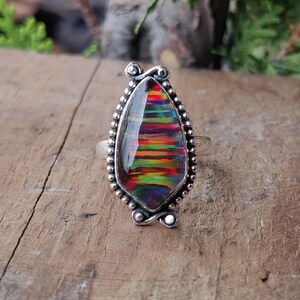 Puede incluir: Un anillo de plata con una piedra preciosa iridiscente en forma de l&aacute;grima. La piedra preciosa muestra una vibrante gama de colores, incluyendo rojo, naranja, amarillo, verde y azul. El anillo tiene un borde detallado con cuentas y detalles decorativos.