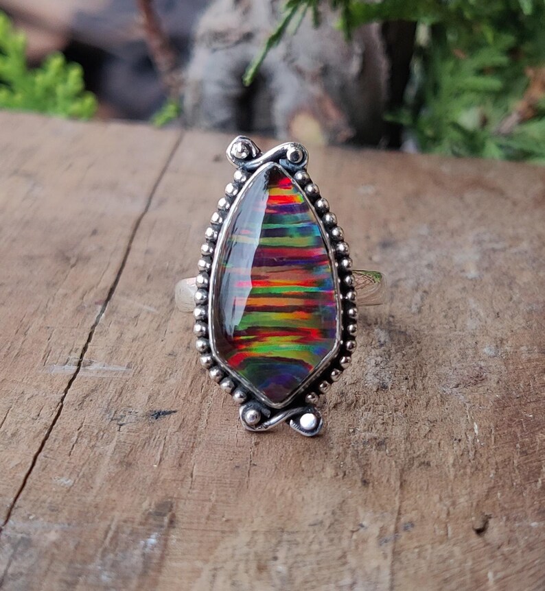 Puede incluir: Un anillo de plata con una piedra preciosa iridiscente en forma de l&aacute;grima. La piedra preciosa muestra una vibrante gama de colores, incluyendo rojo, naranja, amarillo, verde y azul. El anillo tiene un borde detallado con cuentas y detalles decorativos.