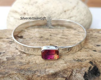 May include: A silver bangle bracelet with a rectangular pink and orange gemstone set in the center.  SilverArtisanGift is printed on the background.