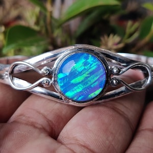 Puede incluir: Una pulsera de plata con una gran piedra de ópalo azul iridiscente y redonda en el centro. La pulsera tiene un diseño calado decorativo con líneas curvas y pequeños acentos redondos. El ópalo muestra tonos de azul y verde.