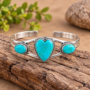 May include: A silver cuff bracelet featuring a central heart-shaped turquoise stone flanked by two smaller oval turquoise stones. Each stone is set within a decorative twisted silver wire border, and the band consists of two parallel silver wires.