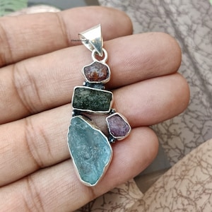 May include: A silver pendant featuring a cluster of five raw gemstones. The pendant is made of sterling silver and features a variety of colors, including green, blue, pink, and red. The pendant is approximately 2 inches in length.