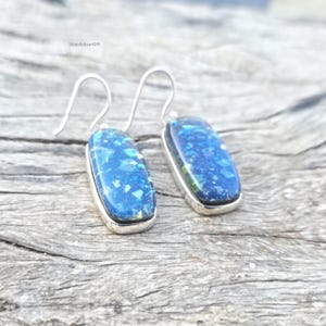 May include: Silver earrings featuring rectangular blue opal stones. The opal stones display a mosaic of blue and green shades, set within a silver bezel. The earrings are suspended from silver hooks.