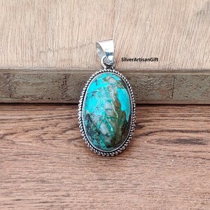 May include: Oval turquoise pendant with silver edging. The turquoise is a vibrant blue-green with white and brown marbling. The pendant is on a wooden surface. SilverArtisanGift is printed in the upper right corner.
