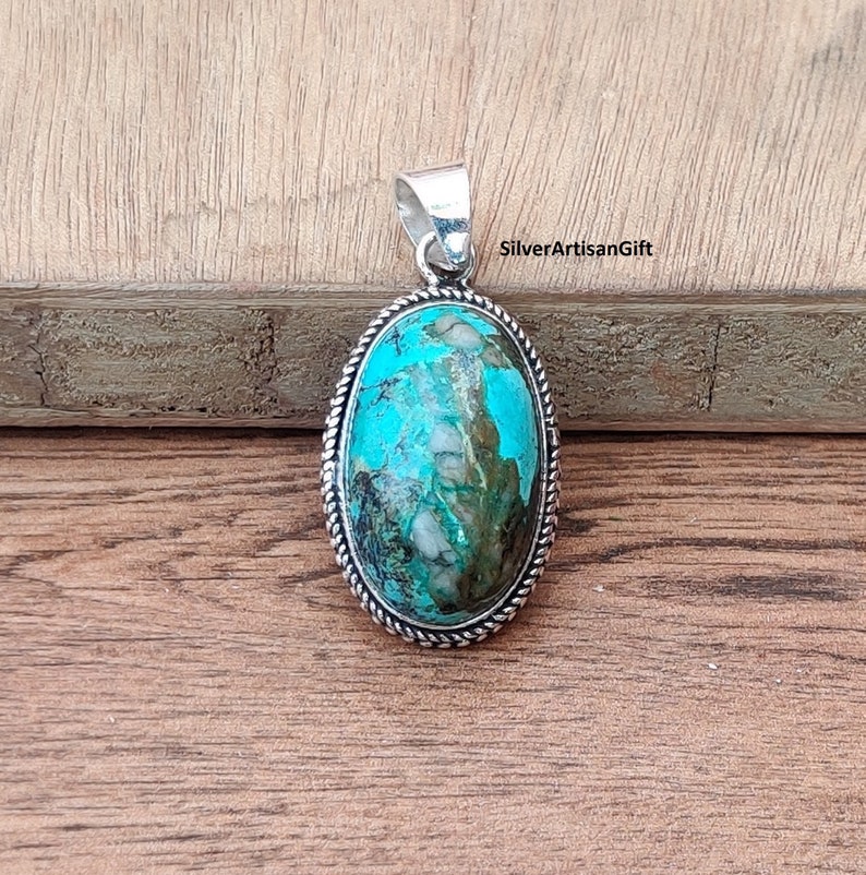 May include: Oval turquoise pendant with silver edging. The turquoise is a vibrant blue-green with white and brown marbling. The pendant is on a wooden surface. SilverArtisanGift is printed in the upper right corner.