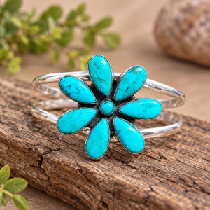 May include: A silver cuff bracelet featuring a central flower motif composed of seven teardrop-shaped turquoise stones with dark matrix veining and a small round turquoise stone at the centre, set against a polished silver metal band structure.