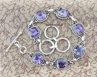 May include: A silver bracelet with a toggle clasp features six square-shaped amethyst gemstones. The bracelet is on a brown background.