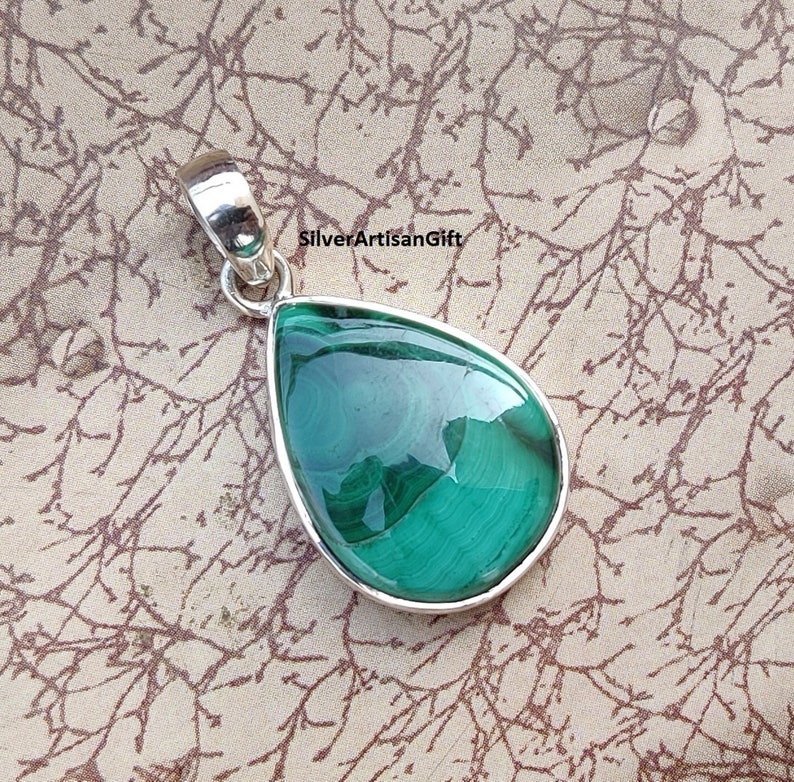 May include: A teardrop-shaped pendant with a polished, vibrant green malachite stone set in a silver bezel. The stone displays natural swirling patterns. The pendant has a silver bail for attaching to a chain. The text "SilverArtisanGift" is visible.