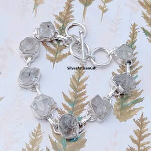 May include: A silver bracelet with a toggle clasp features a row of clear, faceted, rough-cut gemstones.  SilverArtisanGift is printed on the background.
