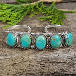 May include: A silver cuff bracelet featuring five oval-shaped turquoise stones set in rope-textured bezels. The metal band displays an engraved pattern along the edges, connecting each stone setting in a continuous, open-ended design.