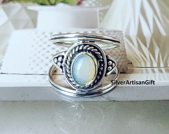 Può includere: Un anello d'argento con una pietra di opale bianca incastonata in una montatura d'argento dettagliata. L'anello è in stile a doppia fascia, con una fascia che avvolge l'altra.