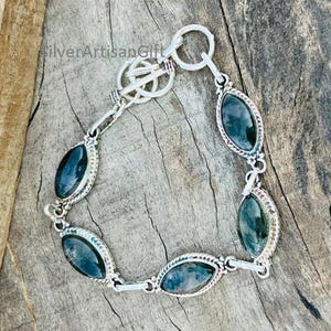 May include: A silver bracelet featuring oval-shaped, dark green gemstones. The gemstones are set in a decorative silver frame. The bracelet has a toggle clasp and is displayed on a wooden surface. The text "SilverArtisanGift" is visible.