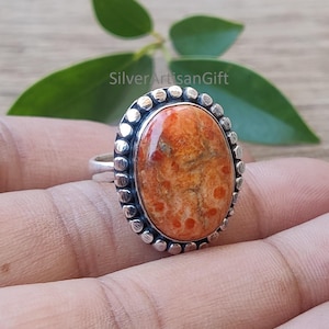 May include: Oval-shaped ring with a silver band and a large, orange-colored stone. The stone has a speckled pattern with red and brown hues. The ring is set against a blurred background of green leaves and wood.