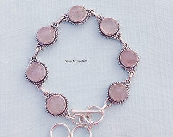 May include: A silver bracelet with a toggle clasp features six round, pale pink gemstones set in silver bezels. The bracelet is made of sterling silver and features a delicate, twisted rope design.