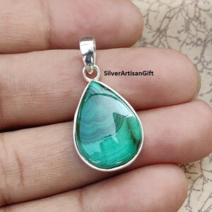 May include: A teardrop-shaped pendant featuring a polished green malachite stone set in a silver-colored bezel. The stone displays natural banding patterns. The pendant has a small loop at the top for a chain.
