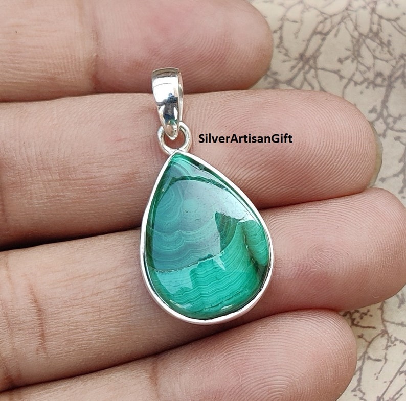 May include: A teardrop-shaped pendant featuring a polished green malachite stone set in a silver-colored bezel. The stone displays natural banding patterns. The pendant has a small loop at the top for a chain.