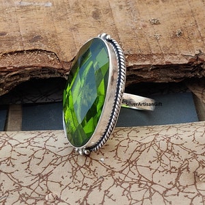 May include: A silver ring with a large, oval-cut, green gemstone. The gemstone is set in a silver bezel with a twisted rope design. The ring has a plain band and the text "SilverArtisanGift". The ring is a piece of jewellery.