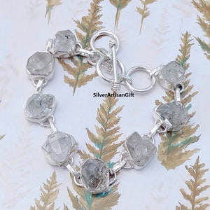 May include: A silver bracelet with a toggle clasp features a row of clear, faceted, rough-cut gemstones.  SilverArtisanGift is printed on the background.
