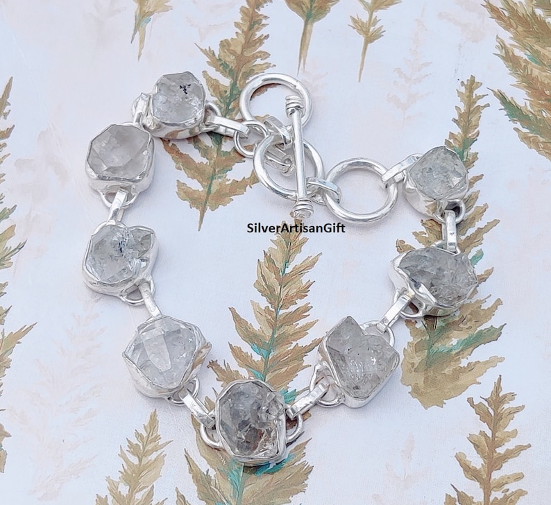 May include: A silver bracelet with a toggle clasp features a row of clear, faceted, rough-cut gemstones.  SilverArtisanGift is printed on the background.