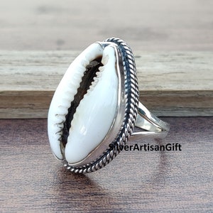 May include: A silver ring with a white cowrie shell set in a rope-like design. The ring is on a wooden surface.