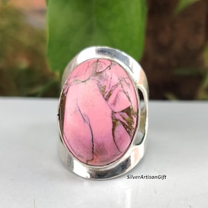Puede incluir: Un anillo de plata con una piedra rosa ovalada grande con vetas marrones y doradas. El anillo tiene una banda ancha con detalles recortados en los lados. El anillo está sobre una superficie blanca con un fondo verde.