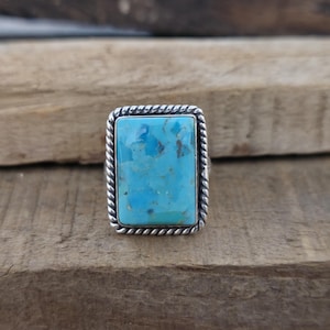 May include: A rectangular turquoise ring with a silver rope-like border. The turquoise stone is a vibrant blue-green color with brown matrix patterns. The ring is set on a wooden surface.
