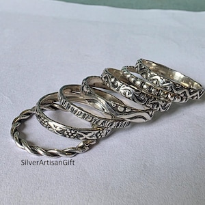 Peut inclure: Plusieurs bagues en argent avec divers motifs, dont des tressés, floraux et géométriques. Les anneaux sont présentés sur une surface de couleur claire. Le texte "SilverArtisanGift" est visible en bas.