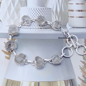 May include: A silver bracelet with a clasp closure features a chain with eight clear, faceted, rough-cut stones. The bracelet is on a white surface with a white and gold patterned surface in the background. SilverArtisanGift is printed in black text on the surface.
