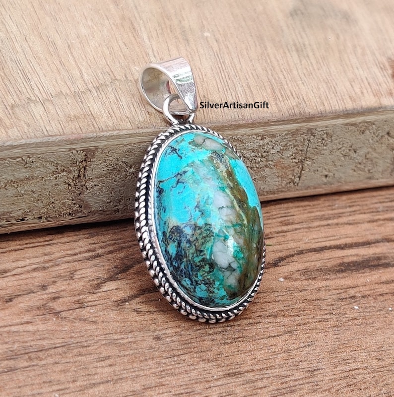 May include: A silver pendant with a turquoise stone set in a silver bezel. The stone has a blue and green color with white and brown markings. The pendant has a rope-like border around the stone.