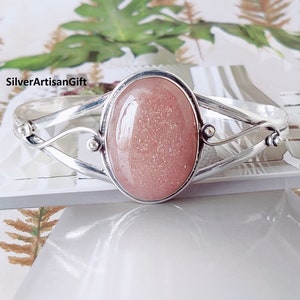 Puede incluir: Un brazalete de plata con un cabujón de piedra de sol rosa. El brazalete tiene un diseño delicado y retorcido.