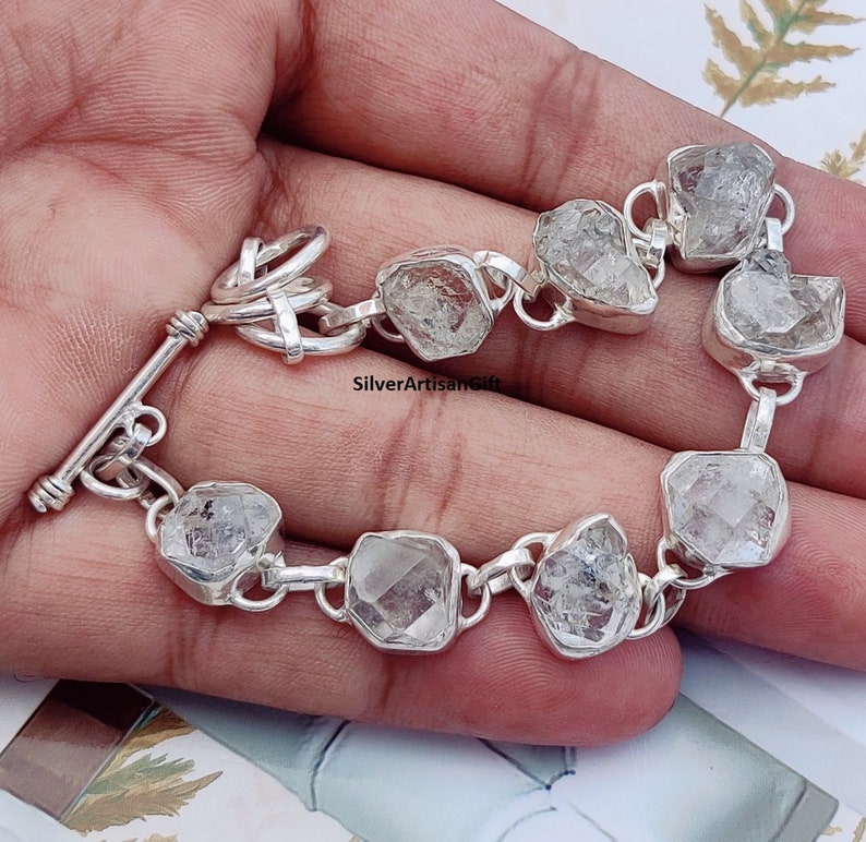 May include: A silver bracelet with a toggle clasp features clear, faceted, rough-cut gemstones.  The bracelet is made of sterling silver and has a polished finish.