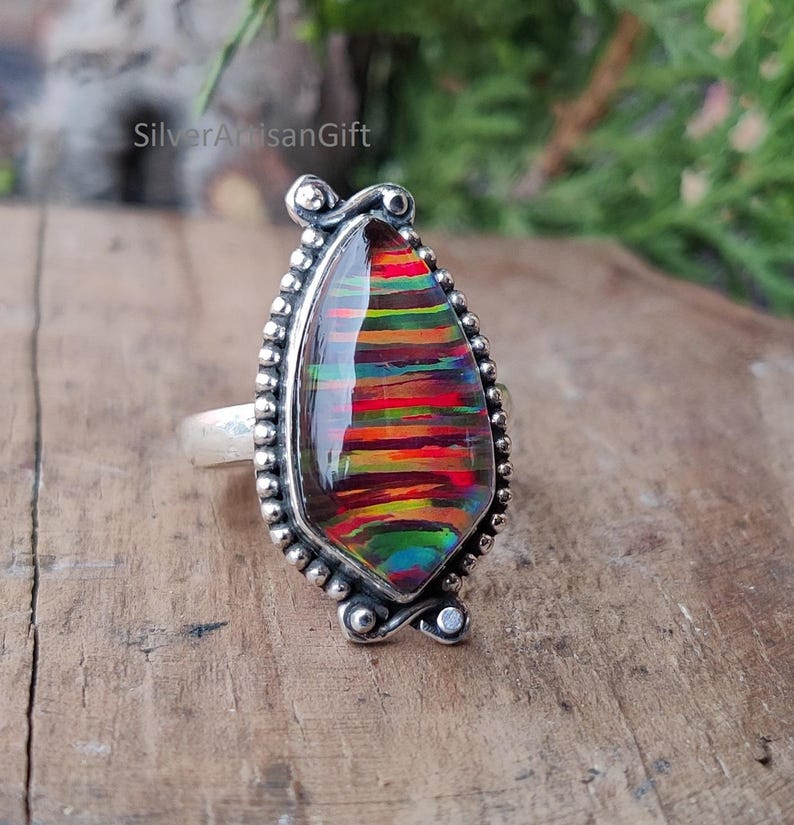 Puede incluir: Un anillo de plata con una gran piedra preciosa en forma de l&aacute;grima. La piedra preciosa muestra una vibrante gama de colores, incluyendo rojo, naranja, amarillo y verde, en un patr&oacute;n de rayas. El anillo tiene un borde detallado con cuentas.