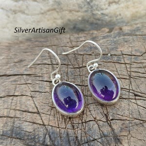 May include: A pair of silver drop earrings with oval, deep purple gemstones. The earrings have a simple design with a hook closure. The text "SilverArtisanGift" is visible in the image.