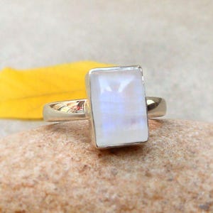 May include: A rectangular moonstone ring with a silver band. The moonstone has a milky white appearance with subtle blue hues. The ring is set on a natural stone surface.