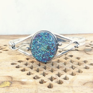 Könnte beinhalten: Ein silbernes Armreif-Armband mit einem großen ovalen, blau-grünen, schillernden Edelstein. Das Armband hat ein zartes, wirbelndes Design.