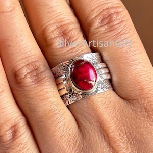 Puede incluir: Un anillo de plata con una piedra ovalada de color rojo intenso. El anillo tiene un diseño grabado y ornamentado alrededor de la banda y un engaste texturizado para la piedra. El anillo está en un dedo.