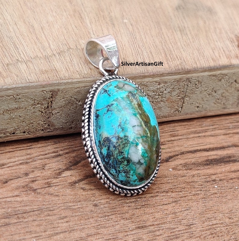 May include: A silver pendant with a turquoise stone set in a silver bezel. The stone has a blue and green color with white and brown markings. The pendant has a rope-like border around the stone.